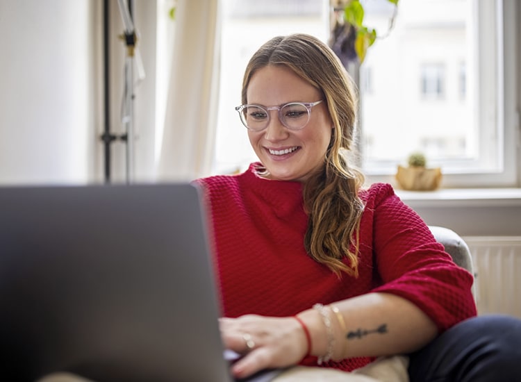 A young woman wearing glasses sitting on a chair while looking at a laptop.
