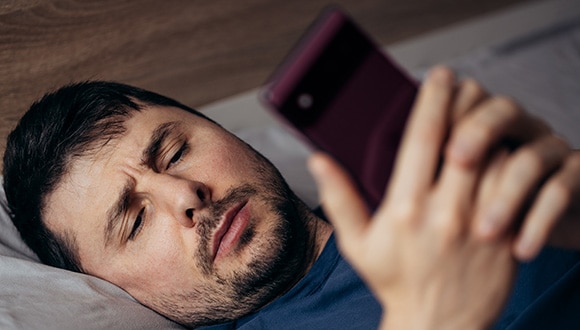 Man laying on his bed while squinting at his phone