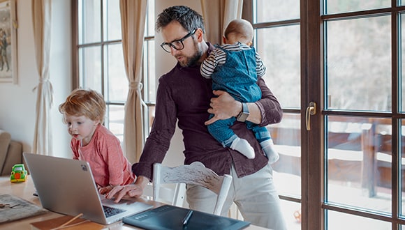 Man on his computer while holding a baby and toddler