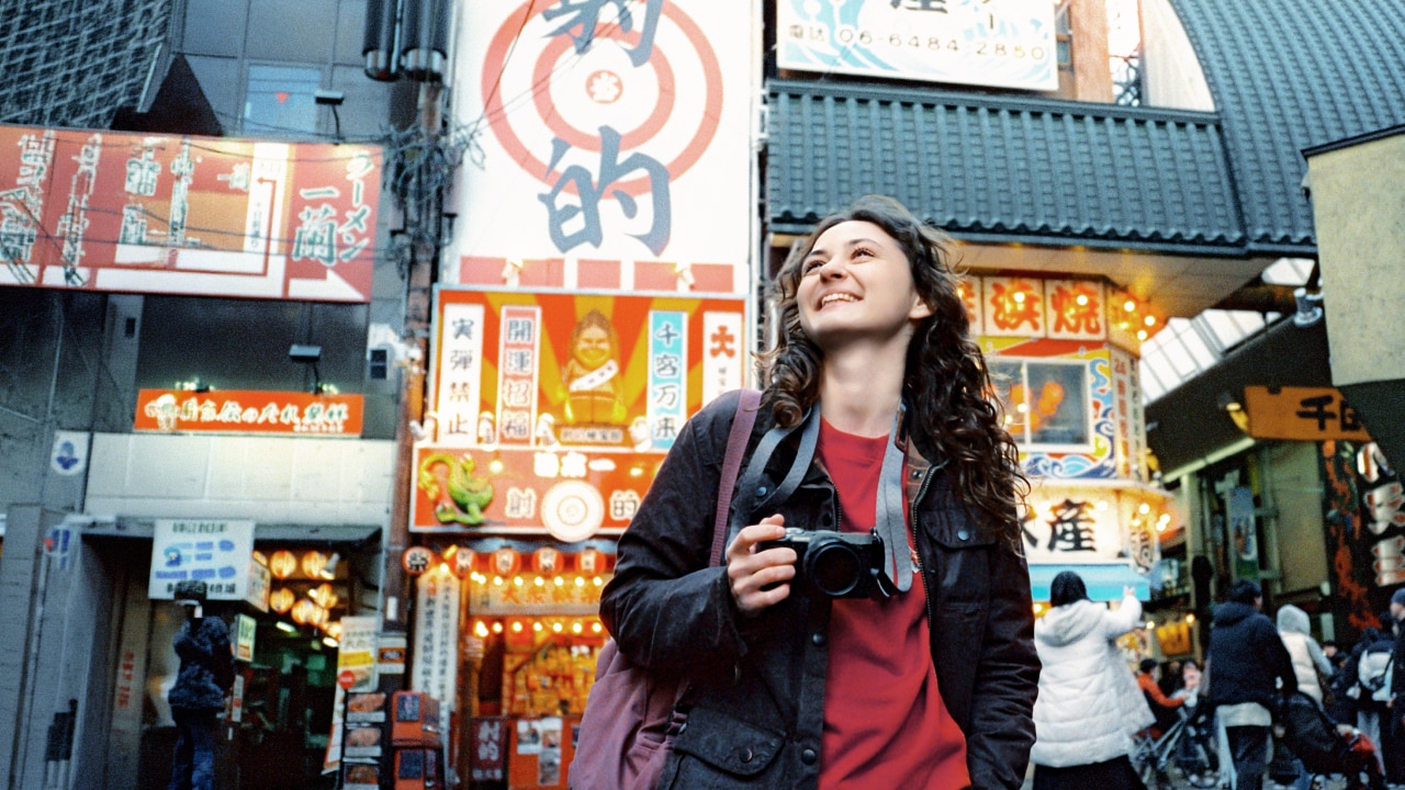 A woman smiling holding a camera with Japanese signage and shops behind her.