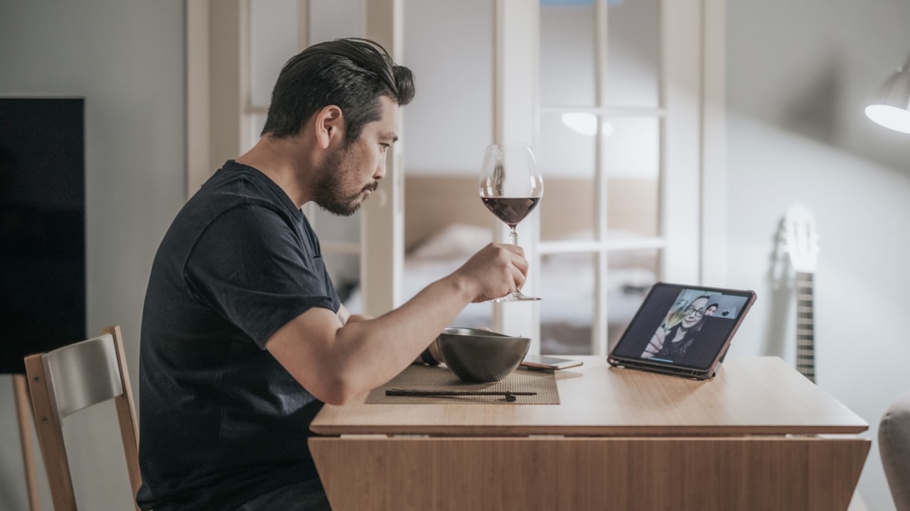 A man holding a glass of wine while on a video call on his tablet while sitting at the dining table.