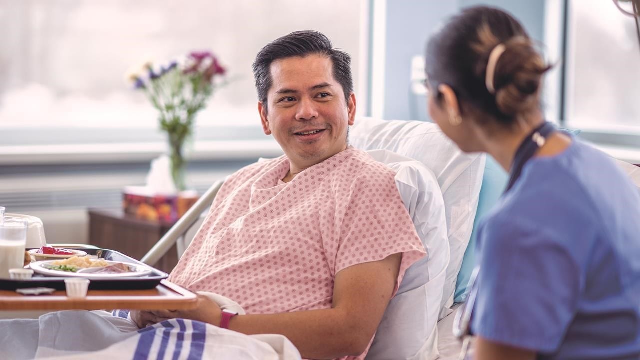 Man laying in hospital bed talking to nurse while eating food.