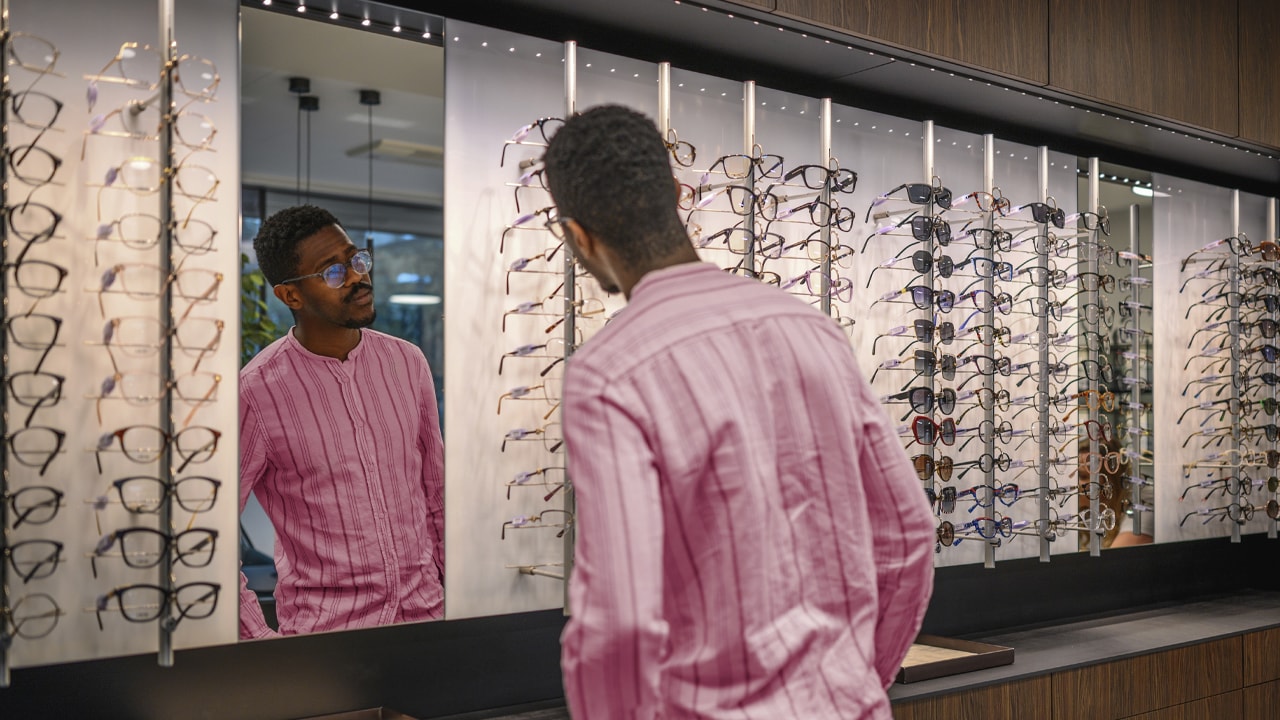 A man facing a wall of glasses looks at his reflection in a mirror while trying some on.