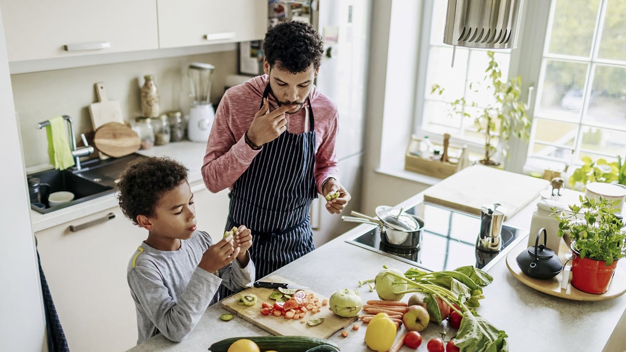A father and son are cooking together in the kitchen and eating some of the ingredients.