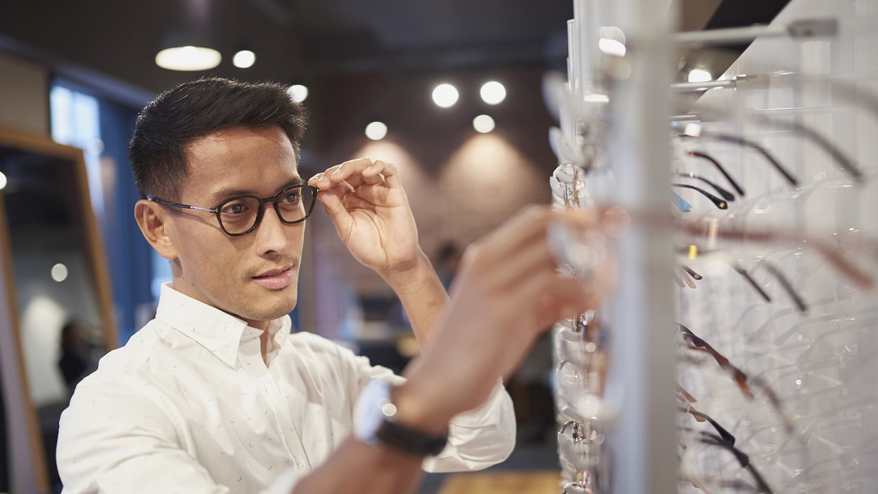 A man tries on a pair of glasses in a store while reaching for another one.