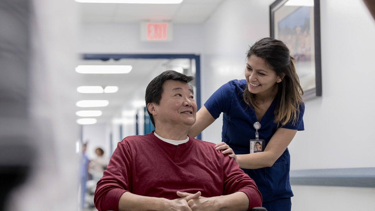 A man sits in a wheelchair in a hospital corridor with a nurse leaning over him and smiling.