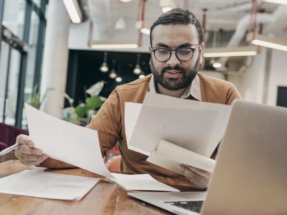 Man looking through a pile of paperwork in front of his laptop.