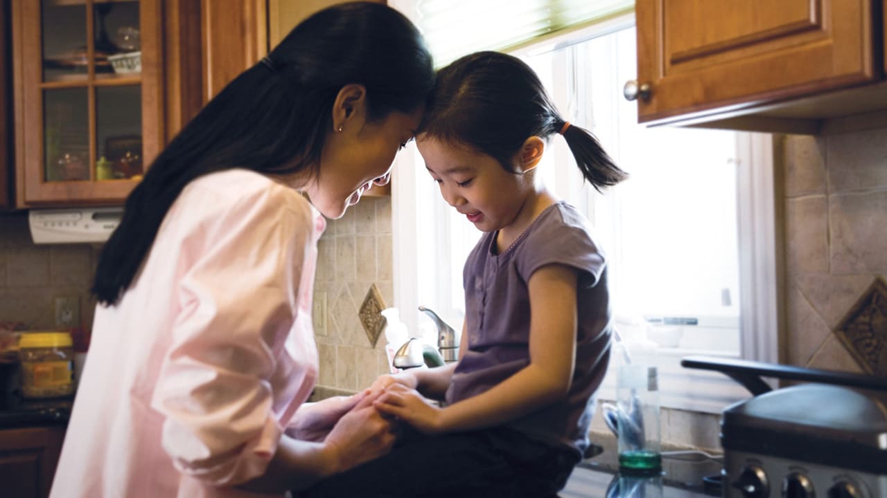A small girl sits on a kitchen bench and her mother holds her hand while their foreheads touch lovingly.