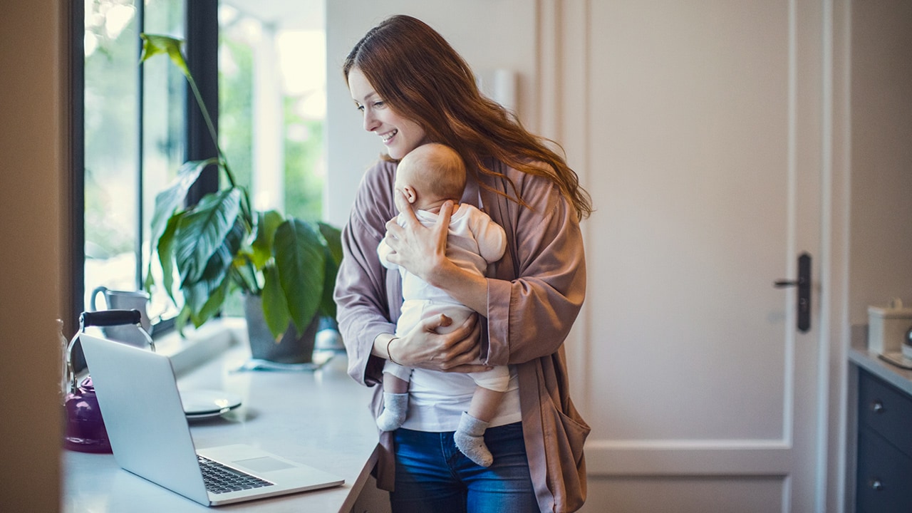 A mother holds her infant child while she looks at a laptop.