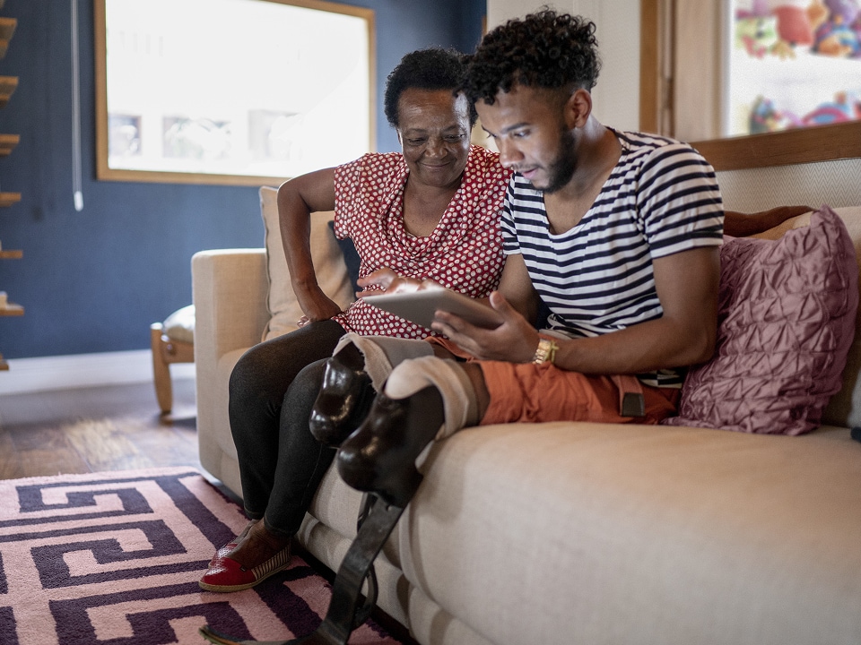 A young man with prosthetic legs, sitting on a couch with a tablet on his lap, showing his grandmother something on the screen.