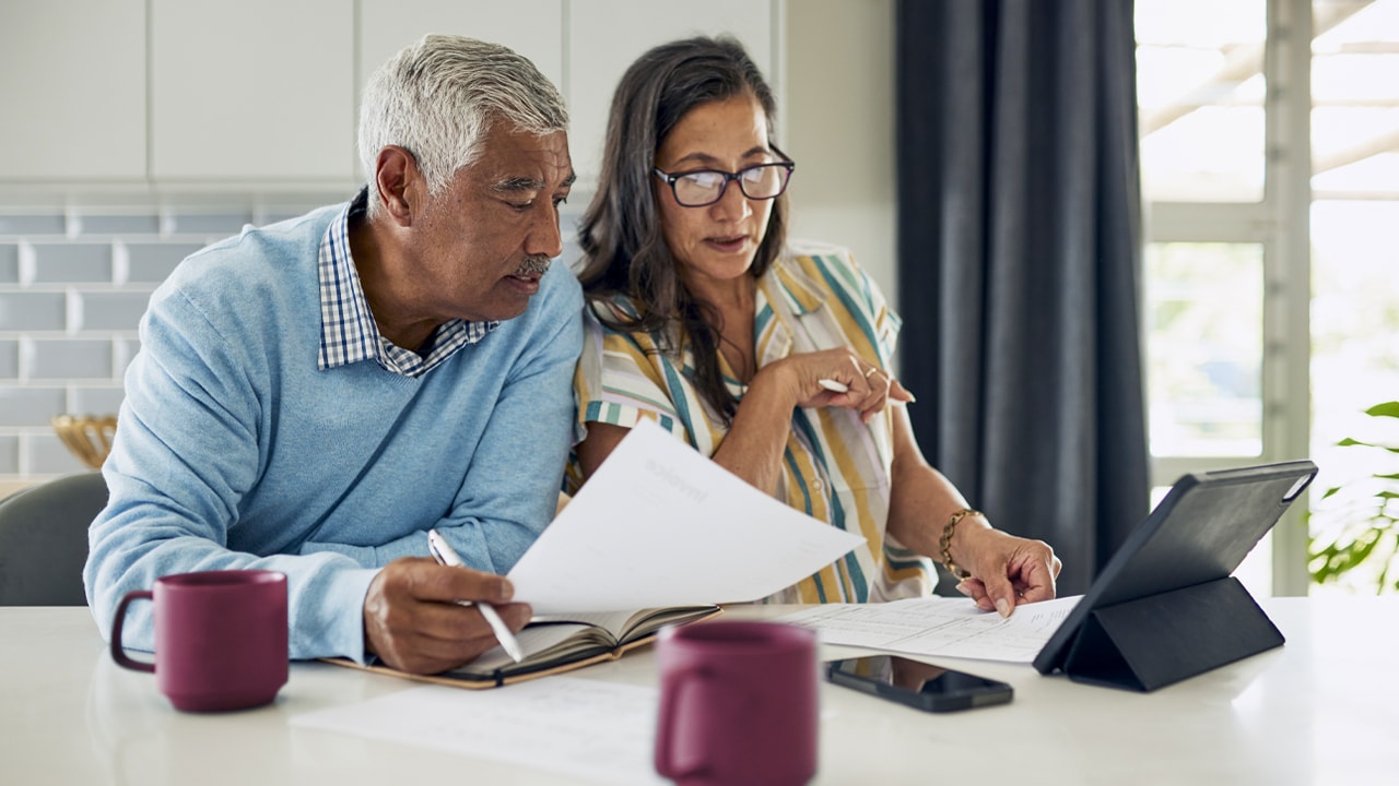 An older couple sitting at the kitchen bench holding documents and looking at a tablet.