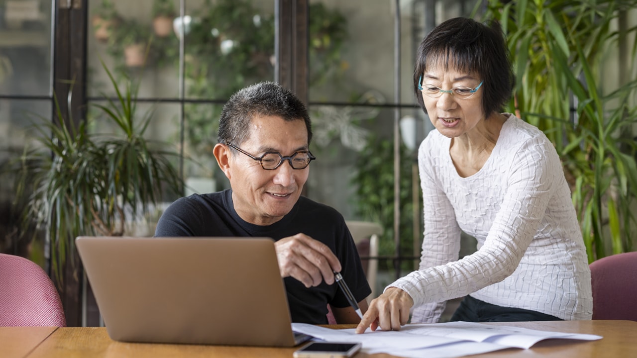 An older man and woman pointing to a document on a table with a laptop in front of them.