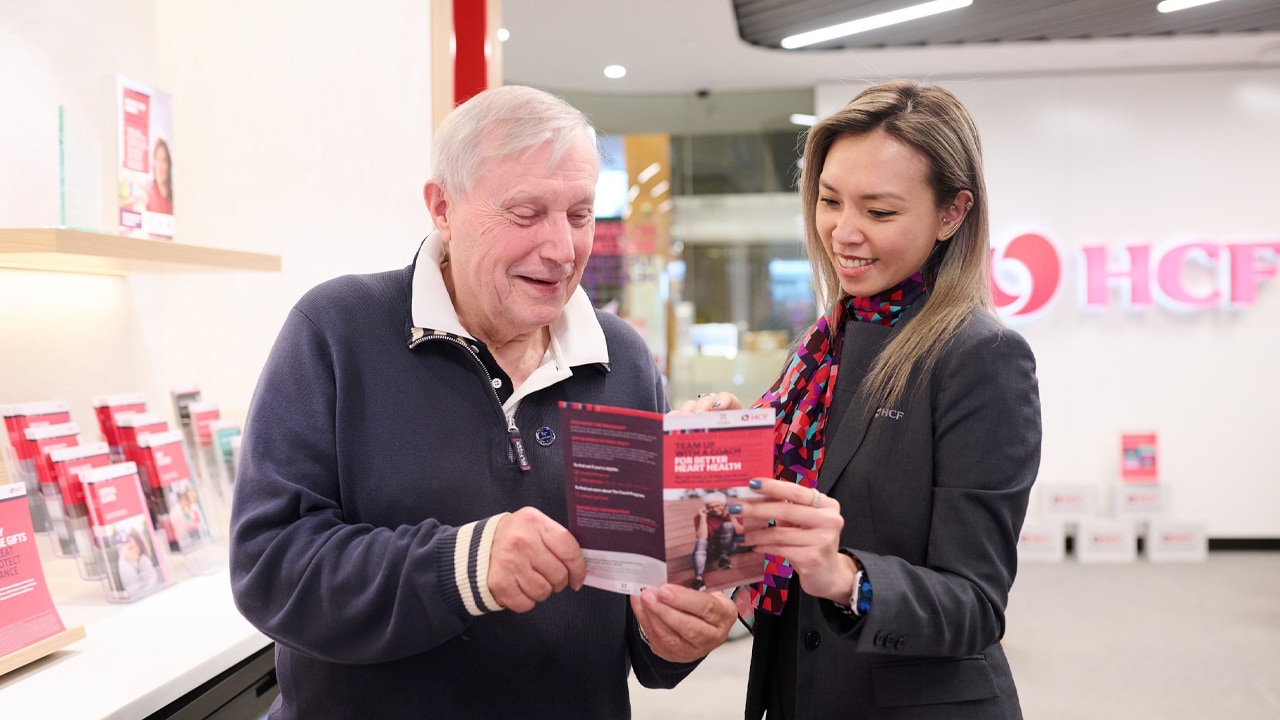 An older man standing next to a HCF staff member while both holding and looking at a health cover brochure.