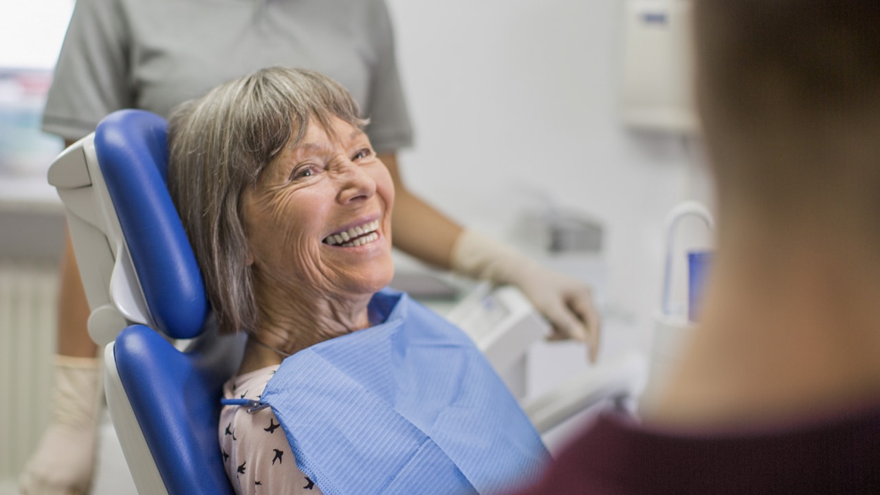 An older woman sits in a dentist's chair smiling with a dentist and assistant in the room.