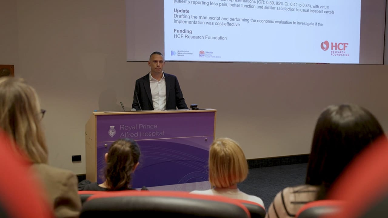 A man stands at a lecturn presenting slides to an audience.