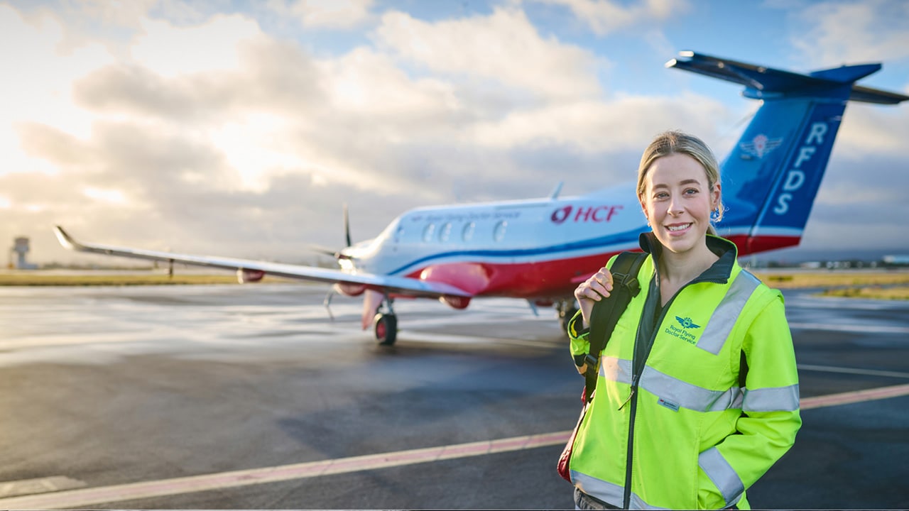Younger woman in high-vis smiles in front a Royal Flying Doctor Service plane with the HCF logo on it