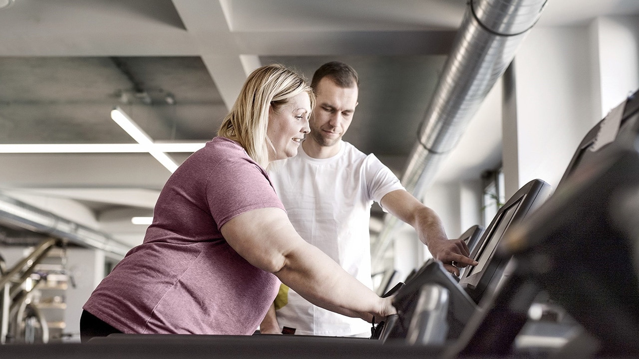 An overweight woman stands on a treadmill and a male personal trainer shows her how to use the display.